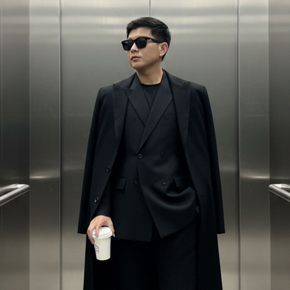 Edit the photo into a leisurely stand inside a modern-style elevator with reflective metal walls.he uses an all-black outfit: an oversize blazer and a long clana using sunglasses, and holding a cup of take-away. elegant and edgy style style with mysterious shades of dim lighting from the ceiling fluorescent lights creates a subtle shadow